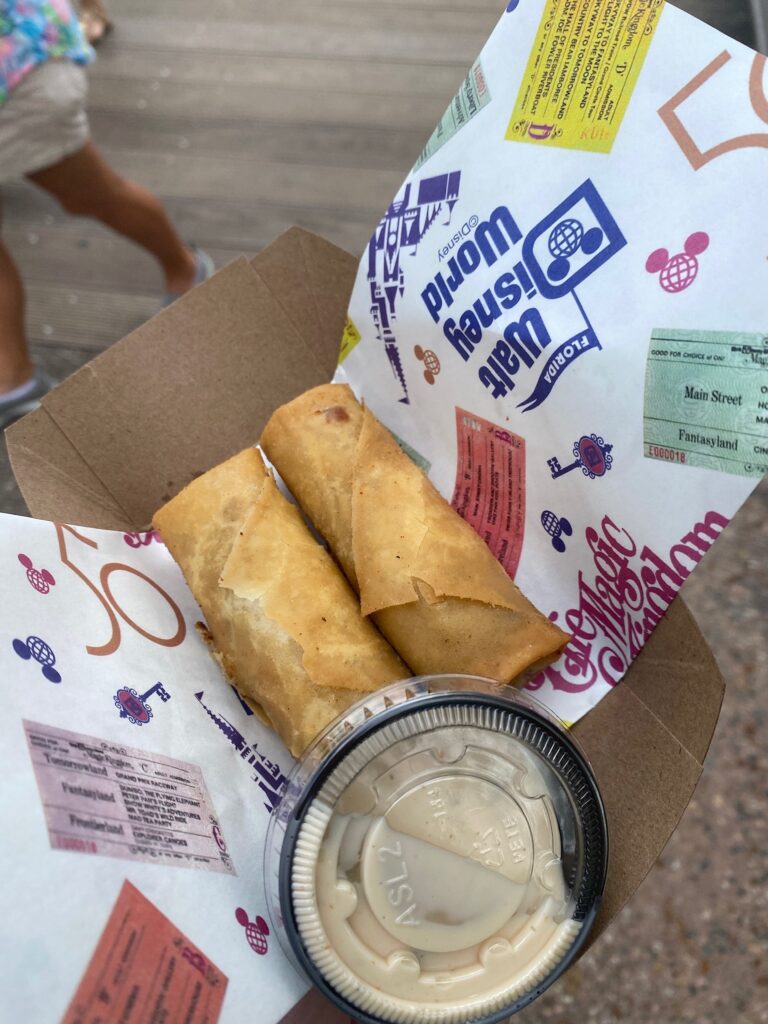 Two fried eggrolls sit in a paper basket with a small container of sauce.
