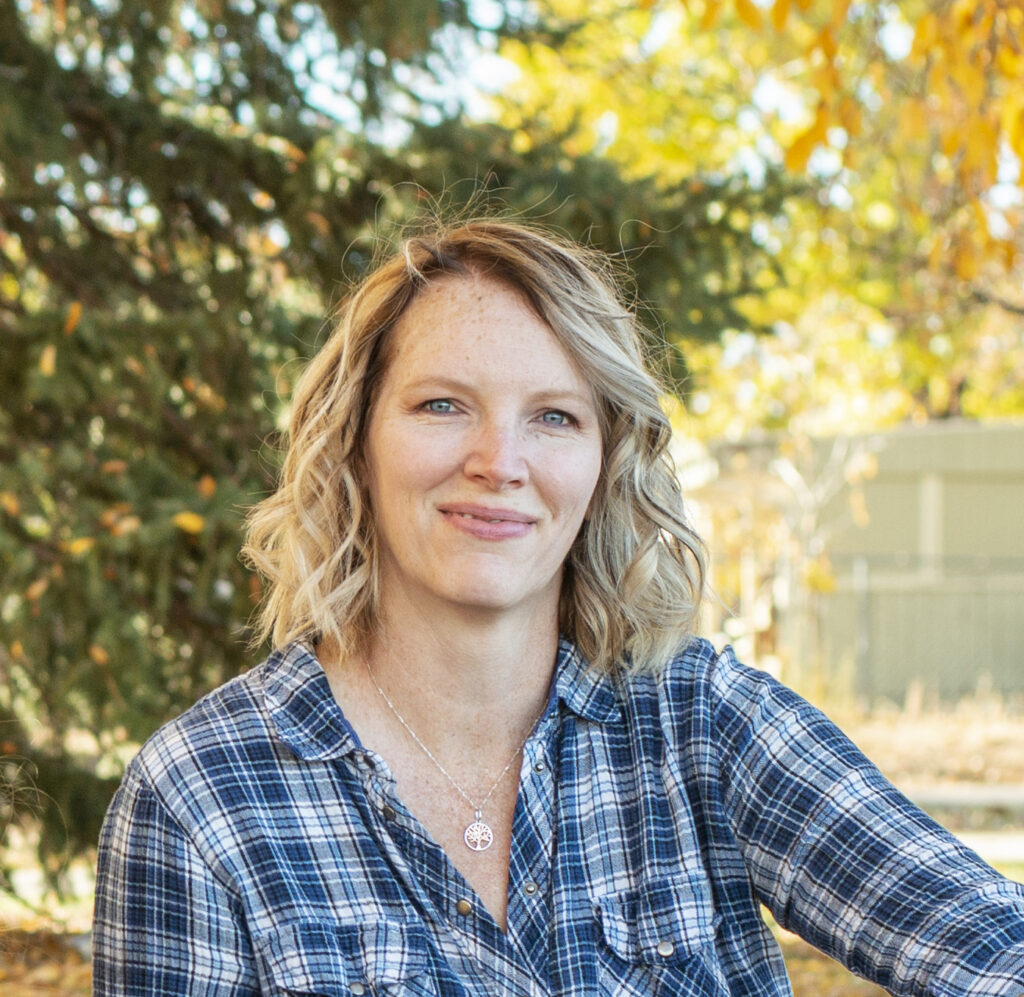 A woman in a blue plaid shirt sits infant of colorful trees in the fall.