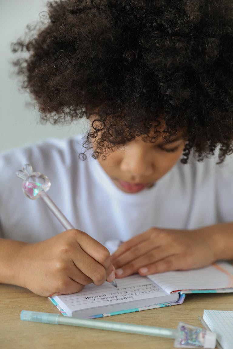 Young African American girl with afro hair writing in a notebook, concentrating on homework indoors.