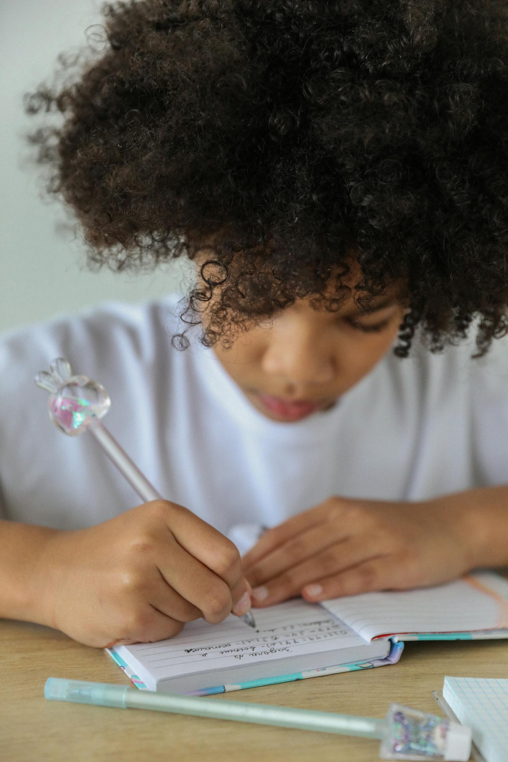 Young African American girl with afro hair writing in a notebook, concentrating on homework indoors.