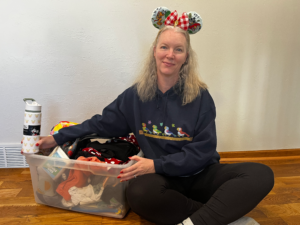 a woman sits next to a plastic bin full of items. She has Disney mouse ears on her head.
