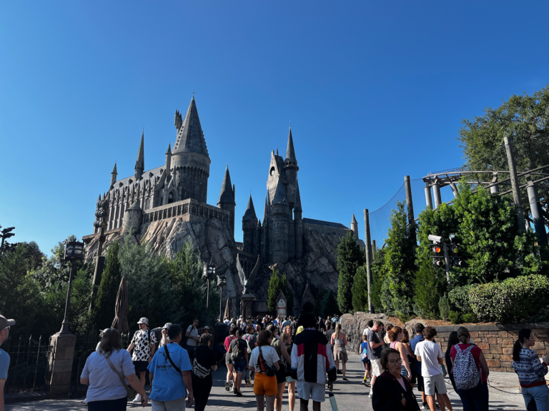 a grey castle stands in the background as a crowd of people walk toward it.