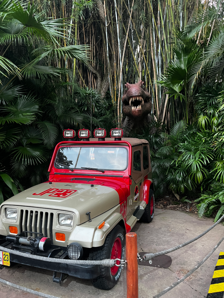 A spinosaurus dinosaur emerges from the trees behind a jeep. 