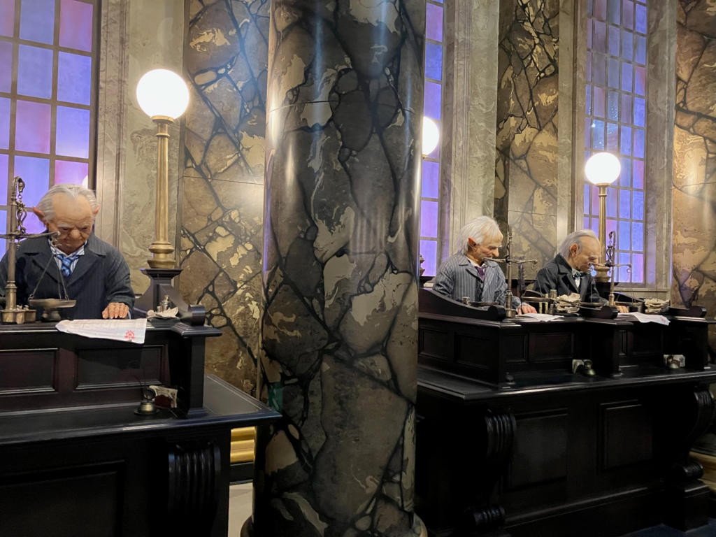 A marble column sits between tall black desks where animatronic goblins appear to work at the wizard bank.