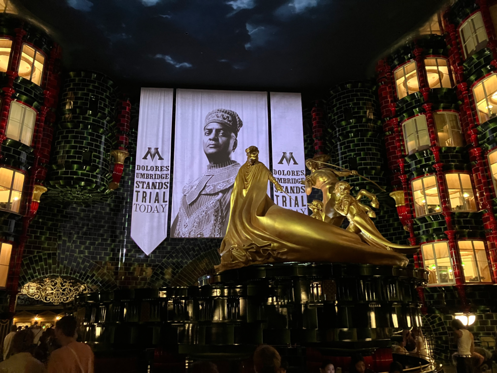  A gold statue of a wizard, a goblin and a centaur sit in the center of a green-tiled room with four floors of office window.