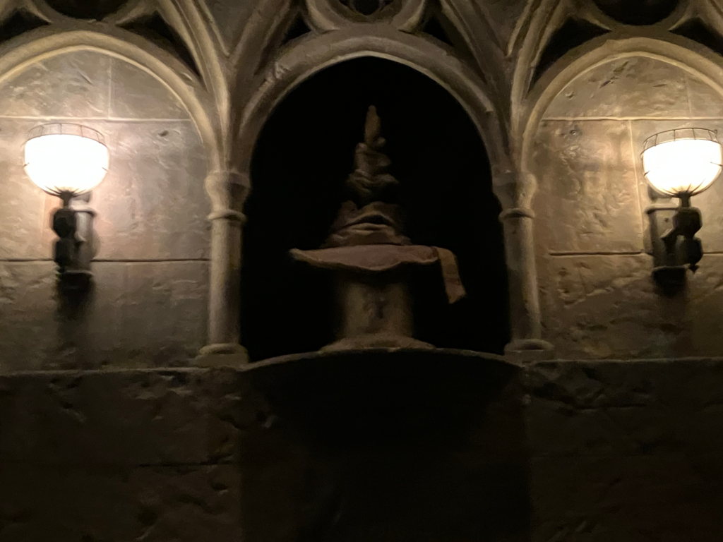 An old pointy hat with a face in the wrinkles sits on a stone pillar in a dark alcove.